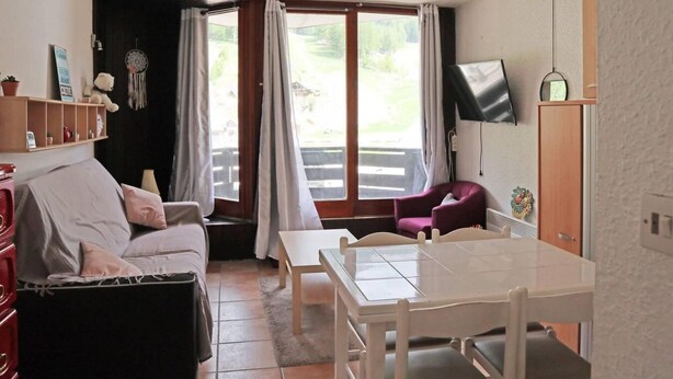 Une pièce confortable avec un canapé, une table à manger et une chaise. Il y a une fenêtre avec des rideaux, un téléviseur accroché au mur et une petite étagère avec quelques objets.
