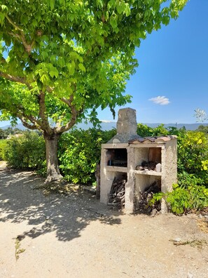 A rustic outdoor grill structure surrounded by greenery and a clear sky.