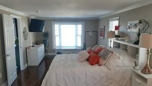A bedroom with a large bed, a dresser, a TV, a window with blinds, and a desk with a lamp.