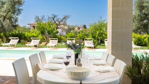 An outdoor dining area with a pool view, white wicker chairs, and a table set for six with wine glasses and a bowl. - Villa La Seta by Perle di Puglia (San Vito dei Normanni)