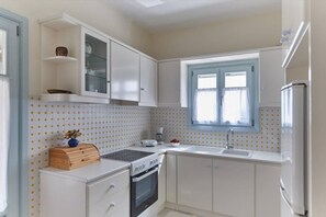 A compact kitchen with white cabinets, a built-in oven, a sink, and a window with curtains. - Aegean Villa in Paros (Paros)