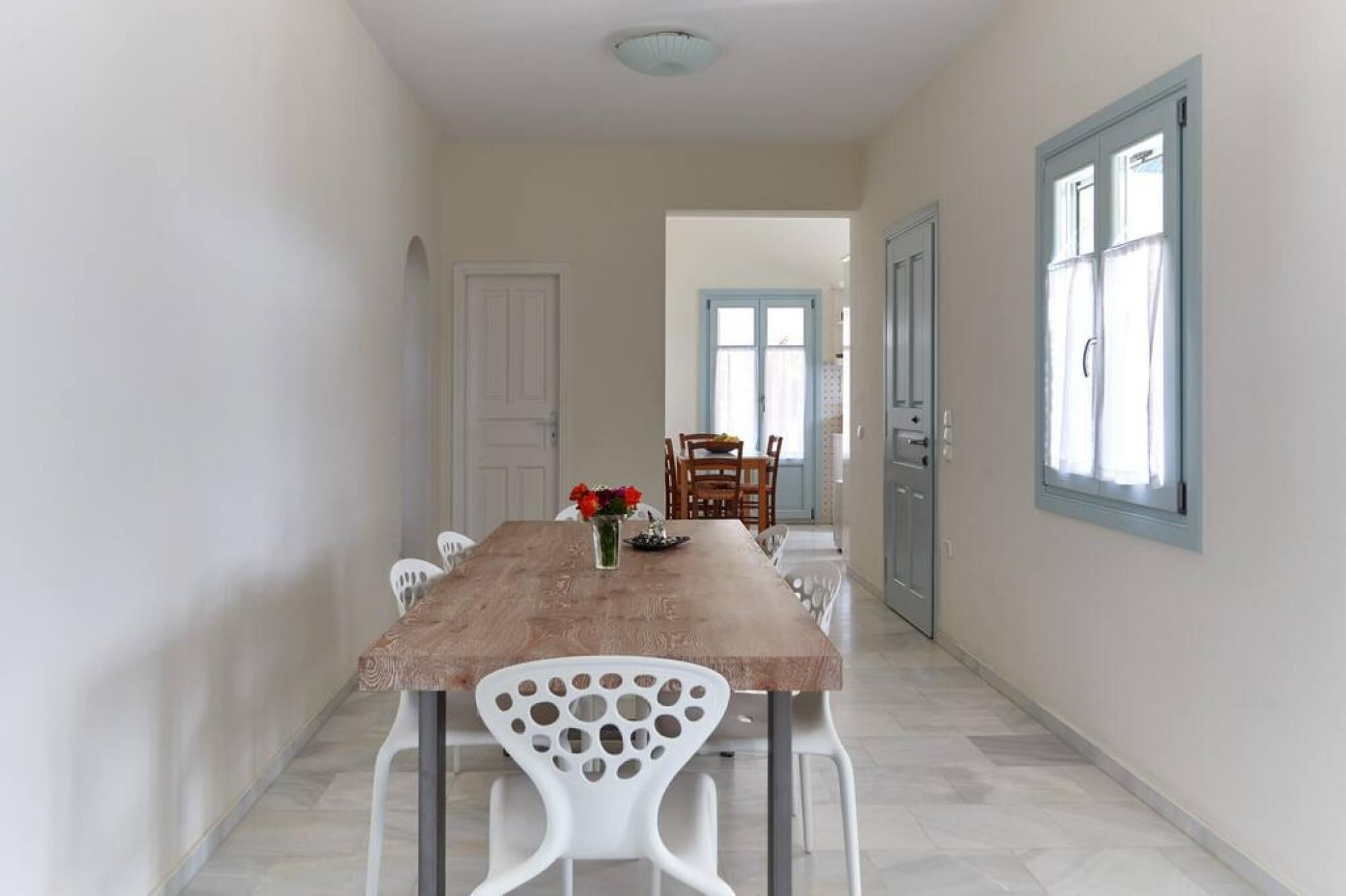 A dining area with a wooden table and white chairs, a vase with flowers, and a view of a blue door and window.