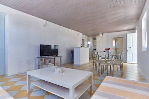 A modern living room with a flat-screen TV, a white coffee table, and a dining area with chairs and a table.