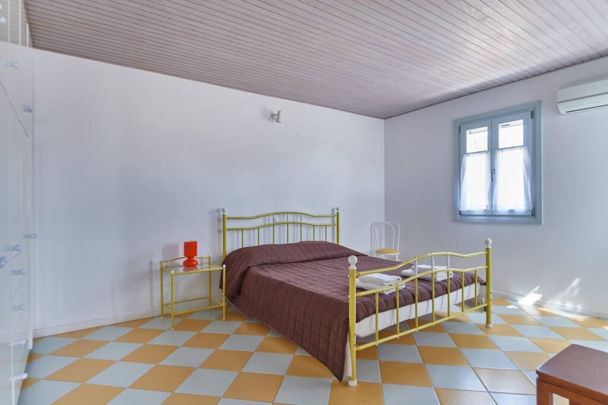 A bedroom with a checkered floor, a bed with a brown bedspread, a nightstand with a red lamp, and a window with curtains.