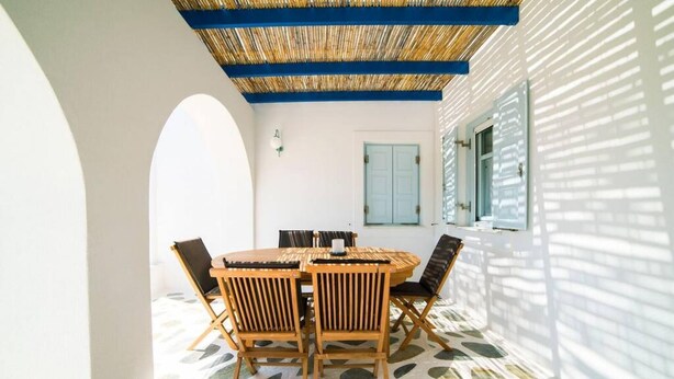 A covered outdoor dining area with a round table and chairs, a patterned floor, and a white building with blue shutters. - Ocean Villa in Paros (Paros)