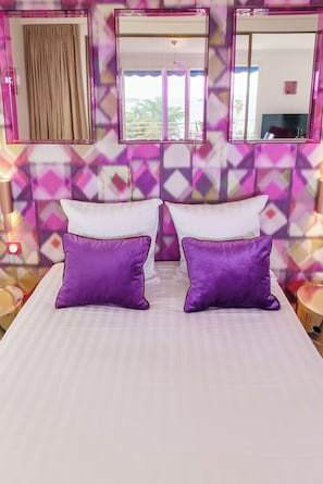 A bed with two white pillows and two purple pillows, a window with a view of palm trees, and a mirror above the bed.