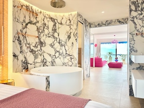 A modern bathroom with a large marble wall, a freestanding bathtub, and a view of a balcony with pink furniture.