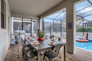 Un patio exterior con una mesa de comedor dispuesta para cuatro personas, rodeada de sillas. Una parrilla es visible a la izquierda, y una piscina con juguetes inflables se ve al fondo.