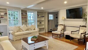 Un salon avec un canapé beige, une table basse en bois et deux fauteuils. Un téléviseur est fixé au mur, et une bibliothèque est remplie de livres et d’objets décoratifs.