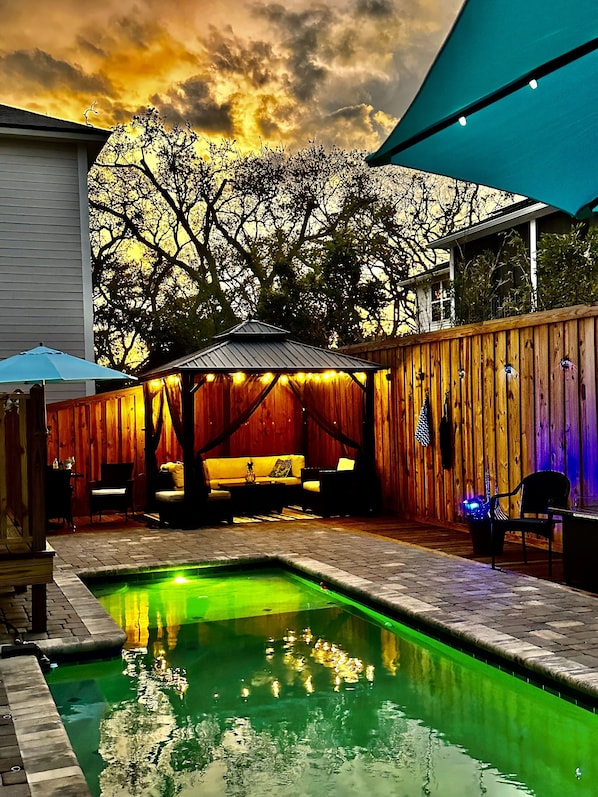 A backyard with a pool, wooden deck, and a covered seating area with string lights.