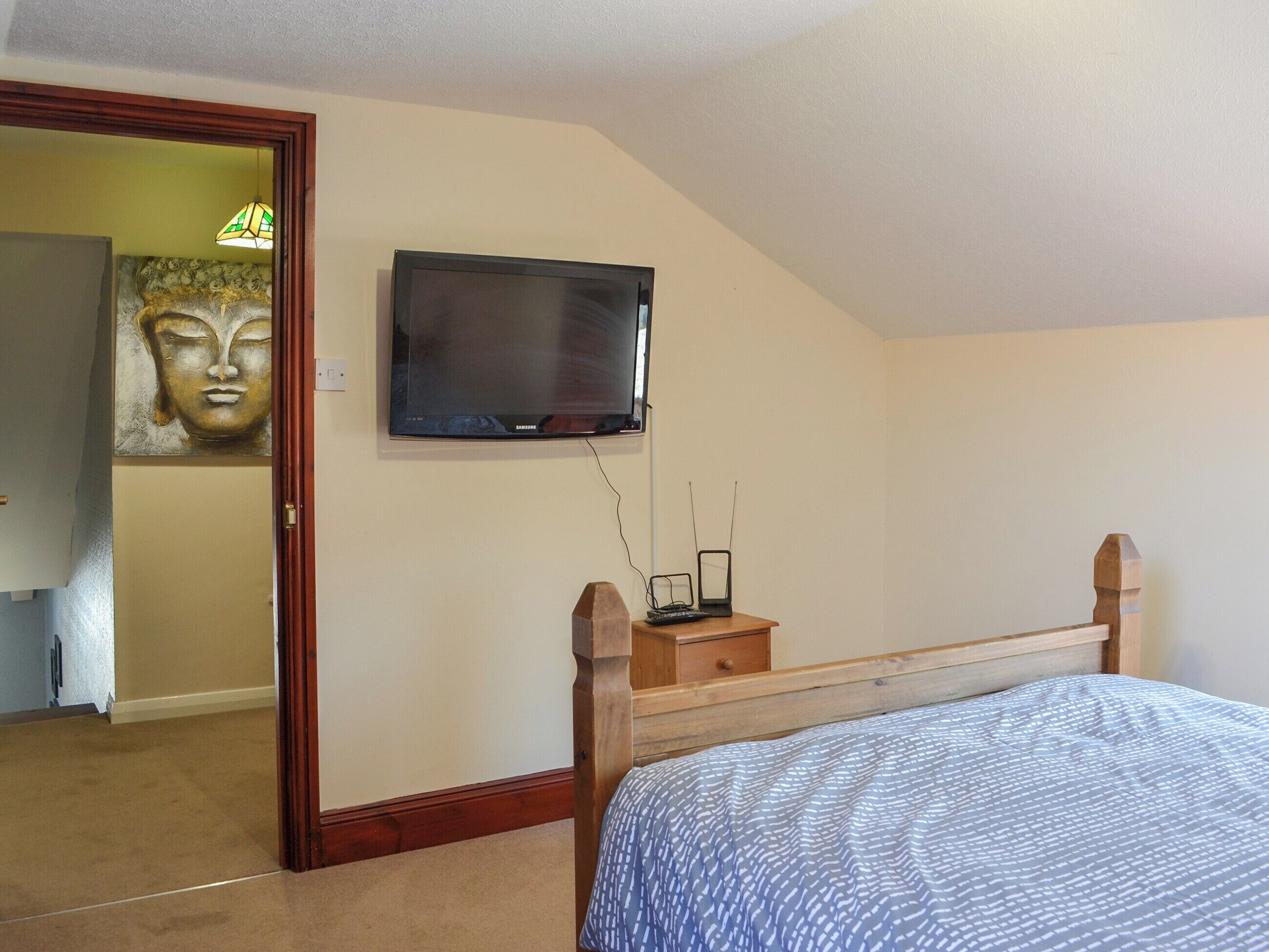 A bedroom with a bed, a television mounted on the wall, a wooden nightstand, and a Buddha statue.
