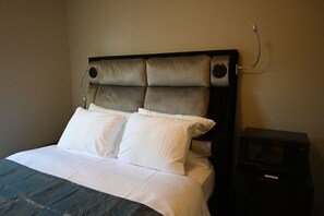 Un lit avec une tête de lit, une literie blanche et une table de chevet noire sur laquelle se trouve un micro-ondes.