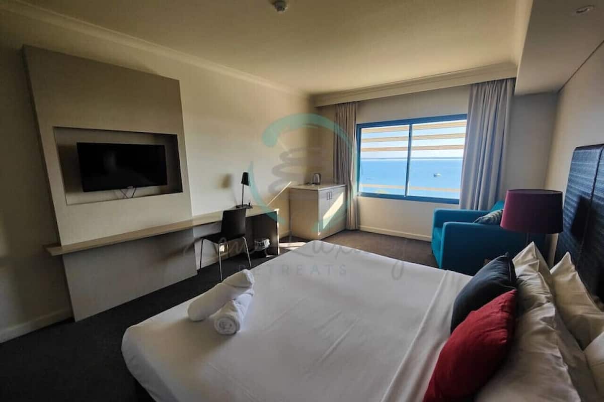 Habitación de hotel con una cama grande, un escritorio, un televisor montado en la pared y vistas al mar desde la ventana.