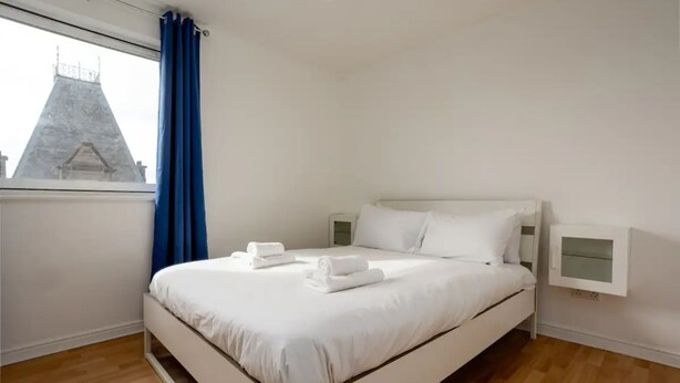 A modern bedroom with a white bed, a nightstand, and a window with blue curtains. - Stylish 2 Bedroom Apartment Near the City Centre (Edinburgh)
