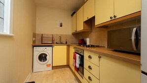 A compact kitchen with a washing machine, oven, microwave, and wooden cabinets. - Stylish 2 Bedroom Apartment Near the City Centre (Edinburgh)