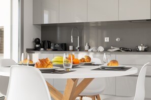 Une cuisine moderne avec des meubles blancs, une table à manger avec des chaises, et un coin petit-déjeuner avec des croissants, des fruits et un verre d’eau.