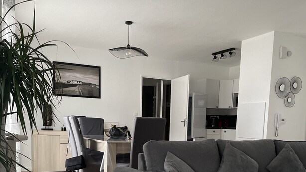 Een moderne woonkamer met een grijze bank, houten vloeren, een eethoek met een tafel en stoelen, en een keuken op de achtergrond.