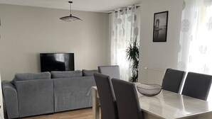 Een moderne woonkamer met een grijze bank, een televisie en een eethoek met een tafel en stoelen.