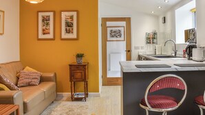 A modern kitchen with a built-in island, a dining area with a red and black chair, and a living room with a sofa and a small table. - 1 bedroom accommodation in Porthcawl (Porthcawl)