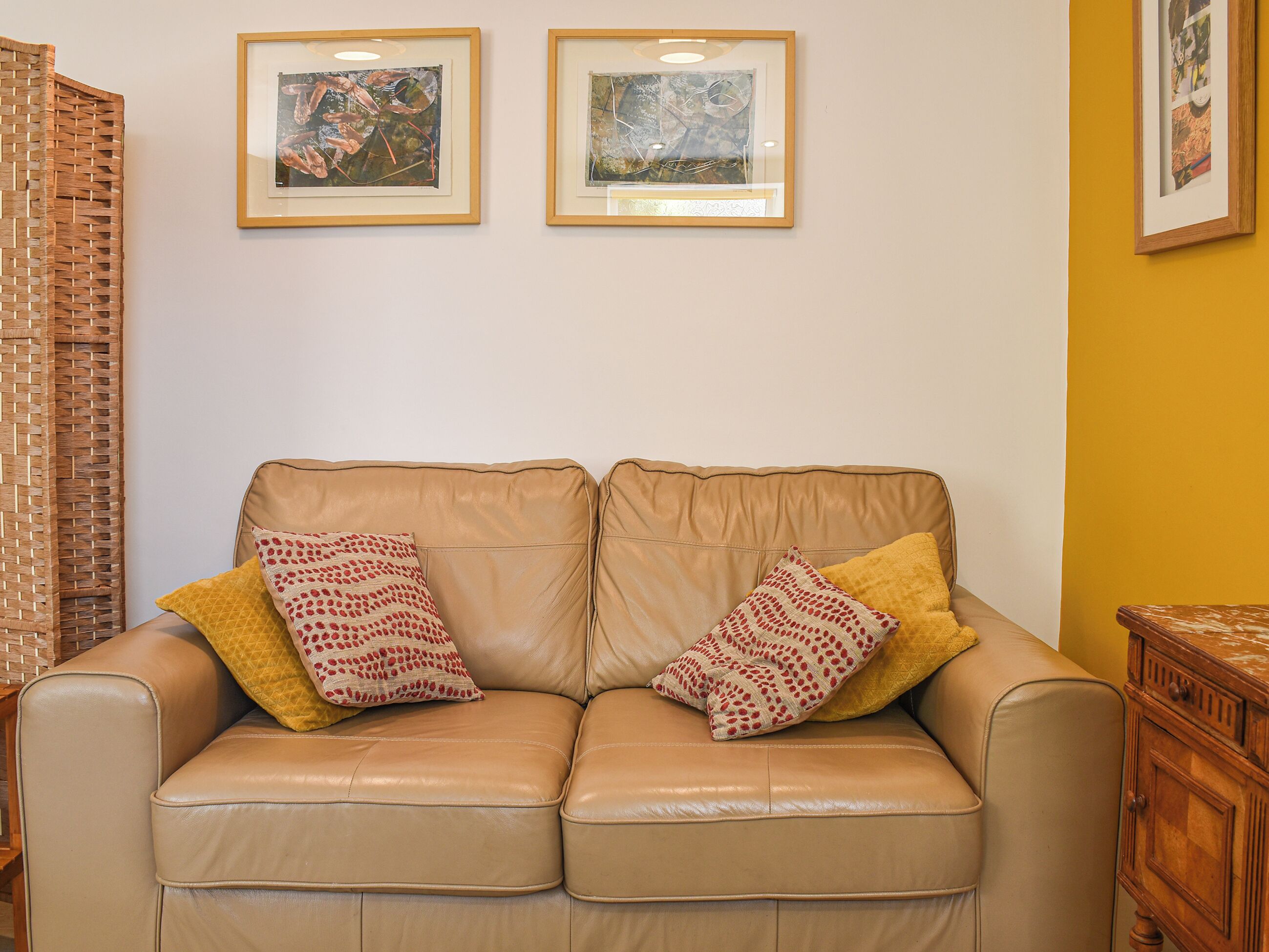 A beige leather sofa with two patterned pillows, a wicker side table, and two framed abstract paintings on the wall.