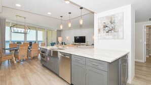 A modern kitchen with grey cabinets, a white countertop, and a dining area with wicker chairs. - Luxury Living from the 9th Floor of Silver Shells (Destin)