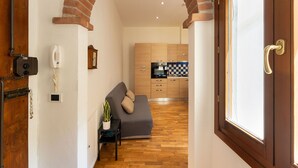 Un couloir mène à une cuisine équipée de meubles en bois, avec un canapé gris et une petite table sur laquelle est posée une plante.
