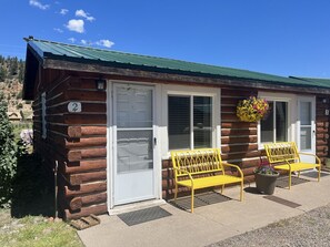 A log cabin with two yellow benches outside, a door with a number, and a hanging flower basket.