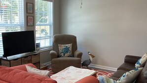 A living room with a ceiling fan, a television on a stand, a brown armchair with a turtle pillow, a red sofa, and a wicker coffee table. - Built in 2021, Cute cottage short walk to beach (Oak Island)