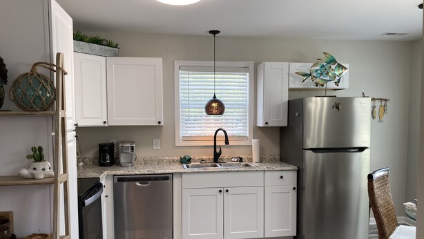 A modern kitchen with white cabinets, stainless steel appliances, a built-in dishwasher, and a window with blinds. - Built in 2021, Cute cottage short walk to beach (Oak Island)