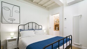 A bedroom with a bed, bedside tables, a wall art piece, a radiator, and a stone wall visible through an open door.
