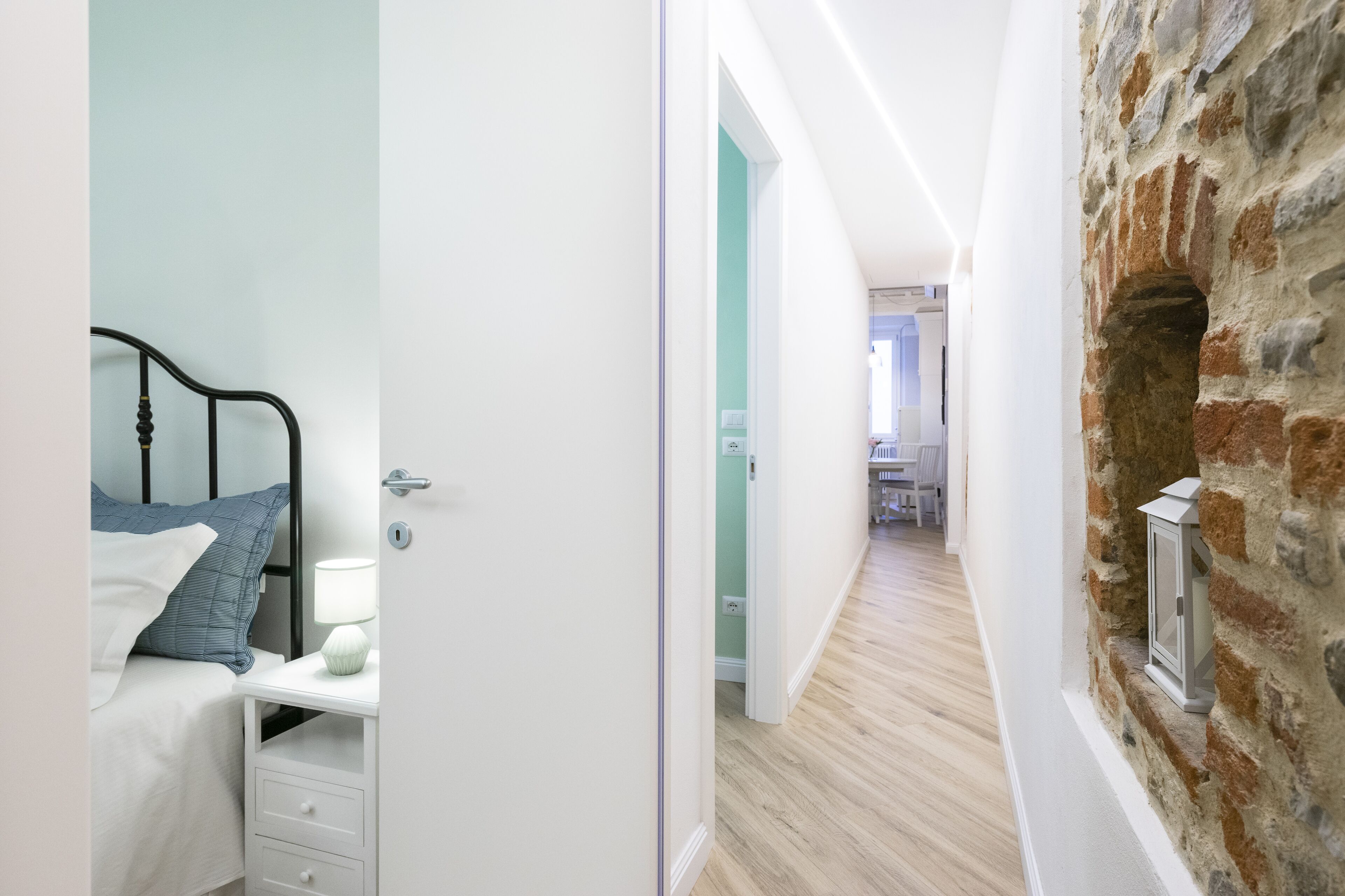 A bedroom with a bed, bedside table, lamp, and a view into a hallway with a stone wall and a door leading to another room.
