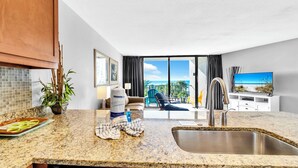 A modern kitchen with granite countertops, a stainless steel sink, and a dishwasher. There is a dining area with a table and chairs, a potted plant, and a view of the ocean through large windows. - 🏖️Stunning Views of Gulf & Pool | Edgewater 1-305🏖️ (Panama City Beach)