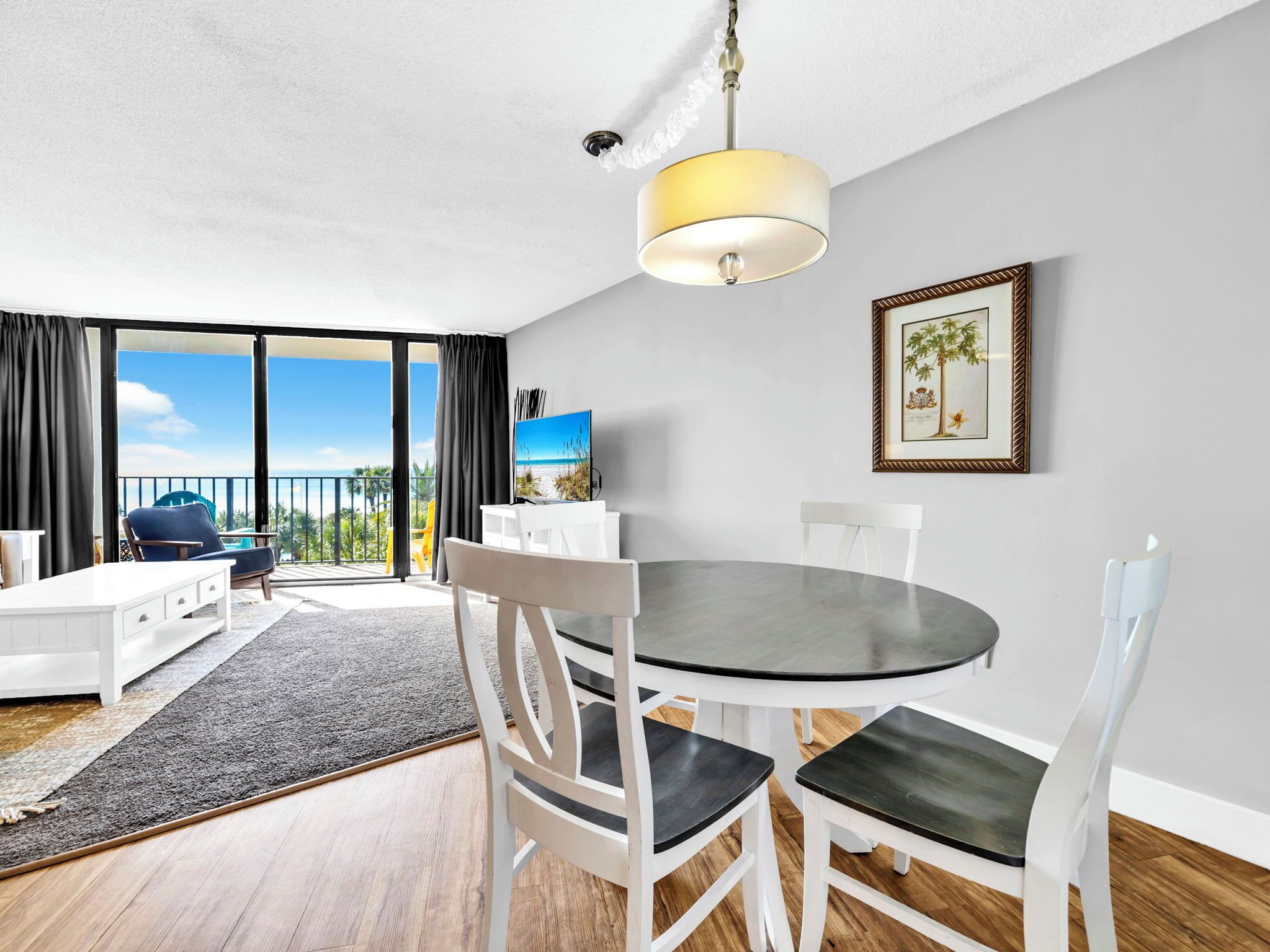 Ein modernes Wohnzimmer mit rundem Tisch, weißen Stühlen, einem Flachbildfernseher und einem großen Fenster mit Blick auf den Strand.