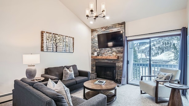 A modern living room with a fireplace, a flat-screen TV, and a large window with a view of snow-covered trees. - Nicely Updated Property, Steps from In-town Shuttle, Pool (Vail)