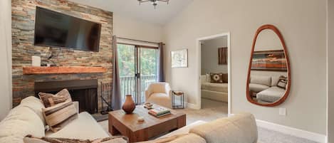 A living room with a stone fireplace, a flat-screen TV, a large mirror, and a view of the outdoors.