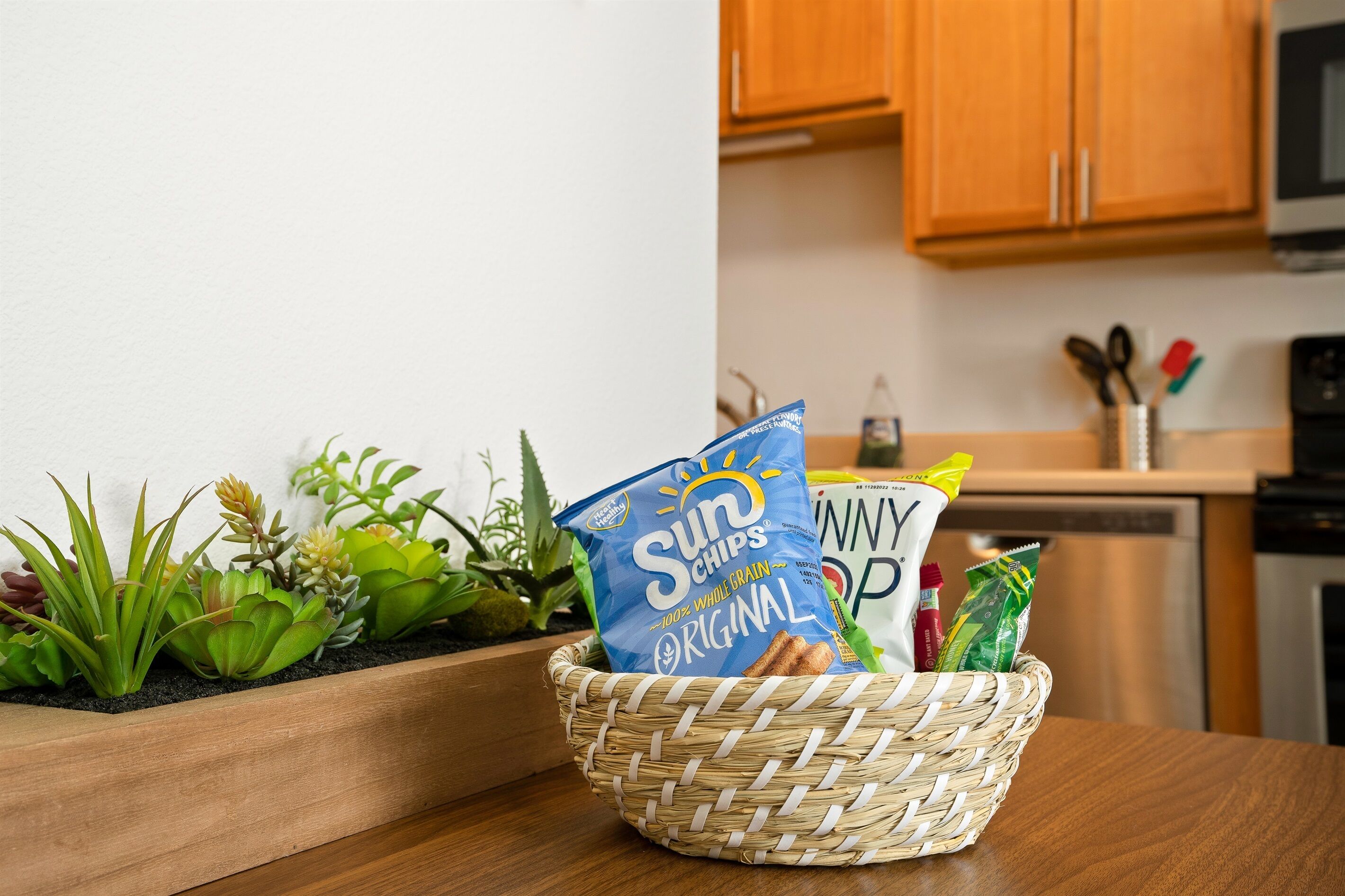A kitchen with wooden cabinets, a countertop, and a woven basket containing snacks and a bottle.