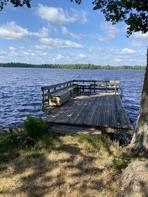 Eine Holzstege in einen See, mit einer Bank und Geländer, umgeben von Bäumen und Rasenflächen.