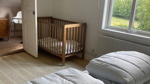 Una habitación con una cuna de madera, una cama con ropa de cama gris, un radiador y una ventana con vistas a zonas verdes.