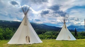 Dos tiendas tipi blancas ubicadas en un campo cubierto de hierba, con montañas al fondo y un cielo nublado.