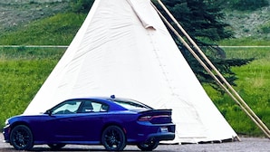 Un coche azul aparcado frente a una estructura blanca similar a un tipi, con una colina boscosa al fondo.