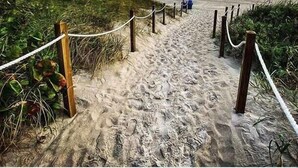 Ein sandiger Strandweg mit hölzernen Pfosten und Seilen, der zum Meer führt.