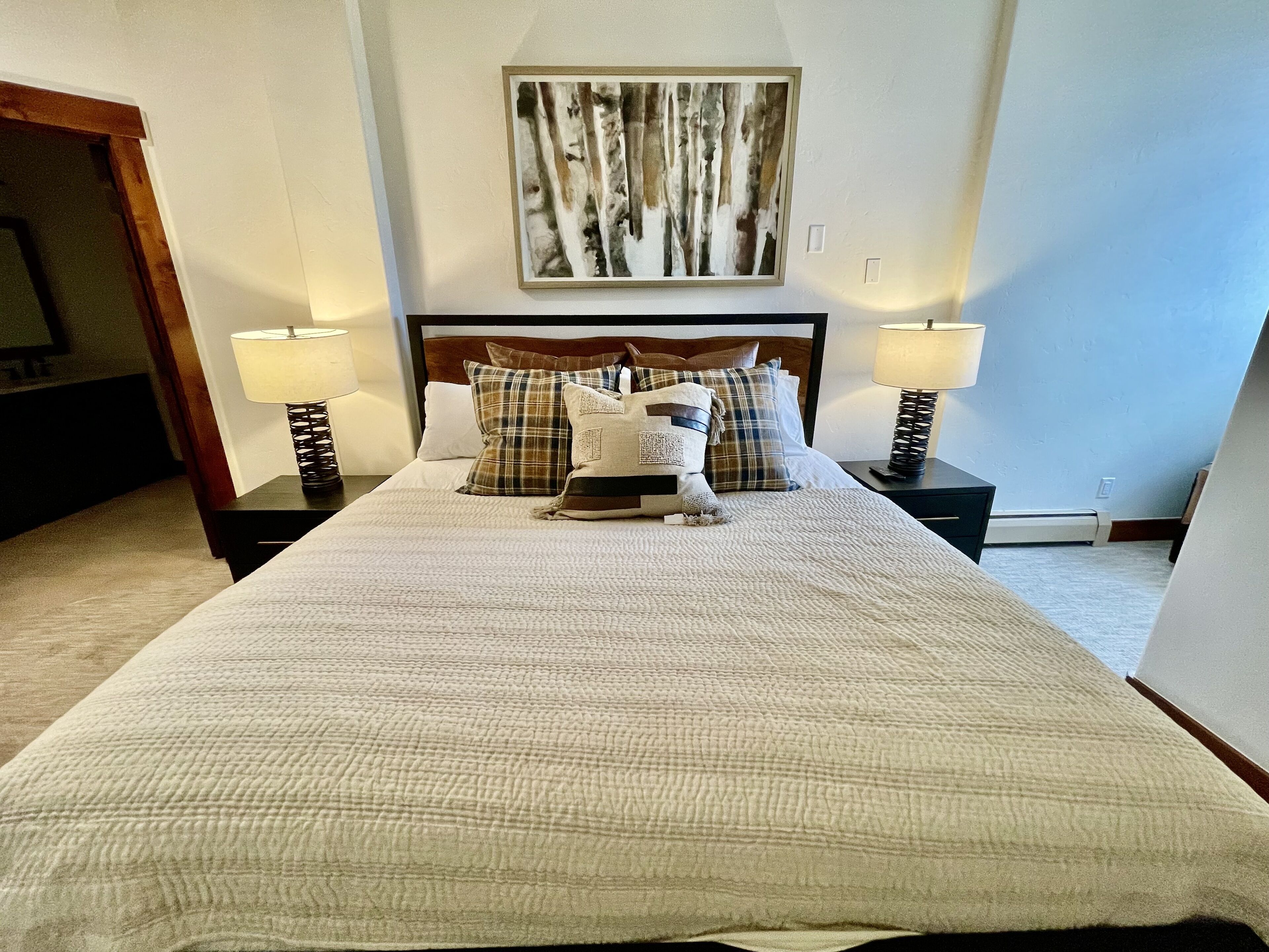 Una cama bien hecha con un edredón blanco texturizado, flanqueada por dos mesitas de noche con lámparas, un cuadro abstracto enmarcado encima y suelo alfombrado.