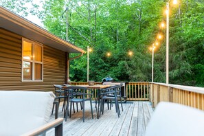 A wooden deck with outdoor furniture and string lights, surrounded by dense forest.