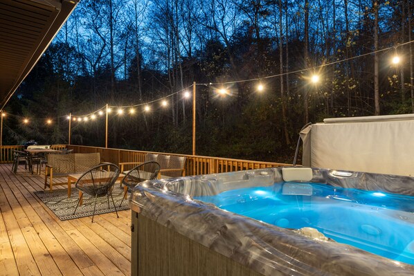 A hot tub on a wooden deck with string lights, surrounded by chairs and a table.