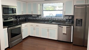 Una cocina con gabinetes blancos, electrodomésticos de acero inoxidable, una encimera de granito y un ventanal con vistas a la vegetación.