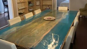 Un comedor con una mesa grande y de textura singular, rodeada de sillas blancas. Sobre la mesa cuelga una lámpara moderna, y se observa una estantería llena de objetos diversos al fondo.