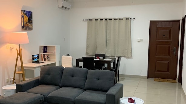 Una sala de estar con sofá modular, un sillón azul, una lámpara, un cuadro, una mesa de comedor con sillas y un ventilador de techo.