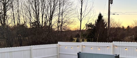 A hot tub in a backyard with a white fence, a wooden utility pole, and string lights.