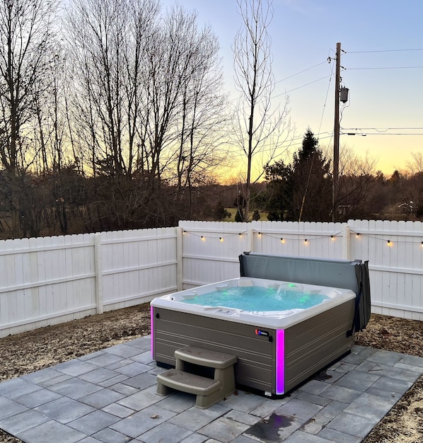 Un jacuzzi en un patio trasero con una cerca blanca, un poste de madera y luces colgantes.