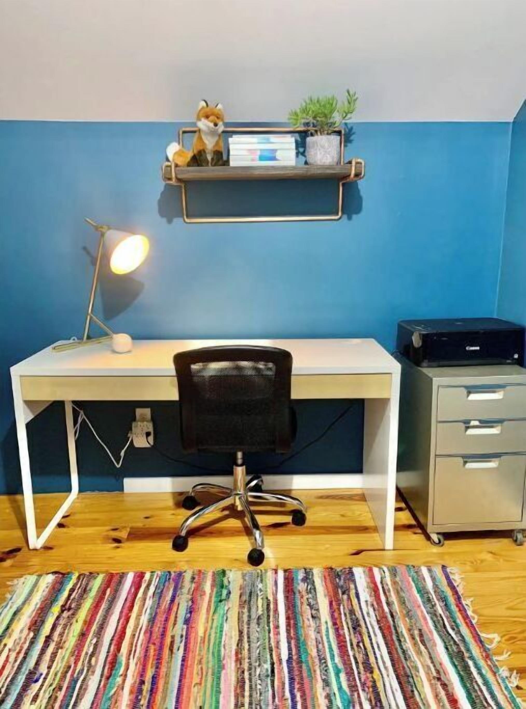 Ein Zimmer mit weißem Schreibtisch, schwarzem Bürostuhl, gestreiftem Teppich, einer Lampe, einem Regal mit einer Fuchsfigur und einem Drucker.